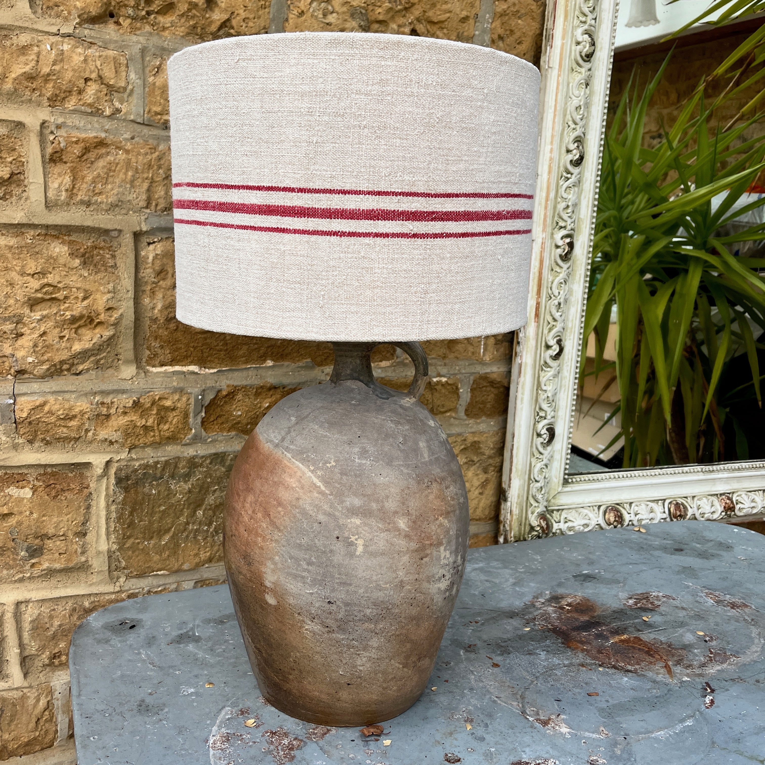 A lamp crafted from a repurposed French olive oil jar, featuring a rustic terracotta base with a narrow neck, topped with a handmade shade made from a Hungarian grain sack. The shade showcases a textured, neutral fabric with subtle vintage patterns