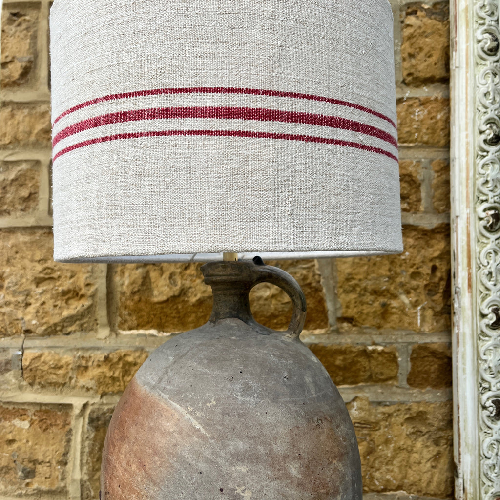 A lamp crafted from a repurposed French olive oil jar, featuring a rustic terracotta base with a narrow neck, topped with a handmade shade made from a Hungarian grain sack. The shade showcases a textured, neutral fabric with subtle vintage patterns