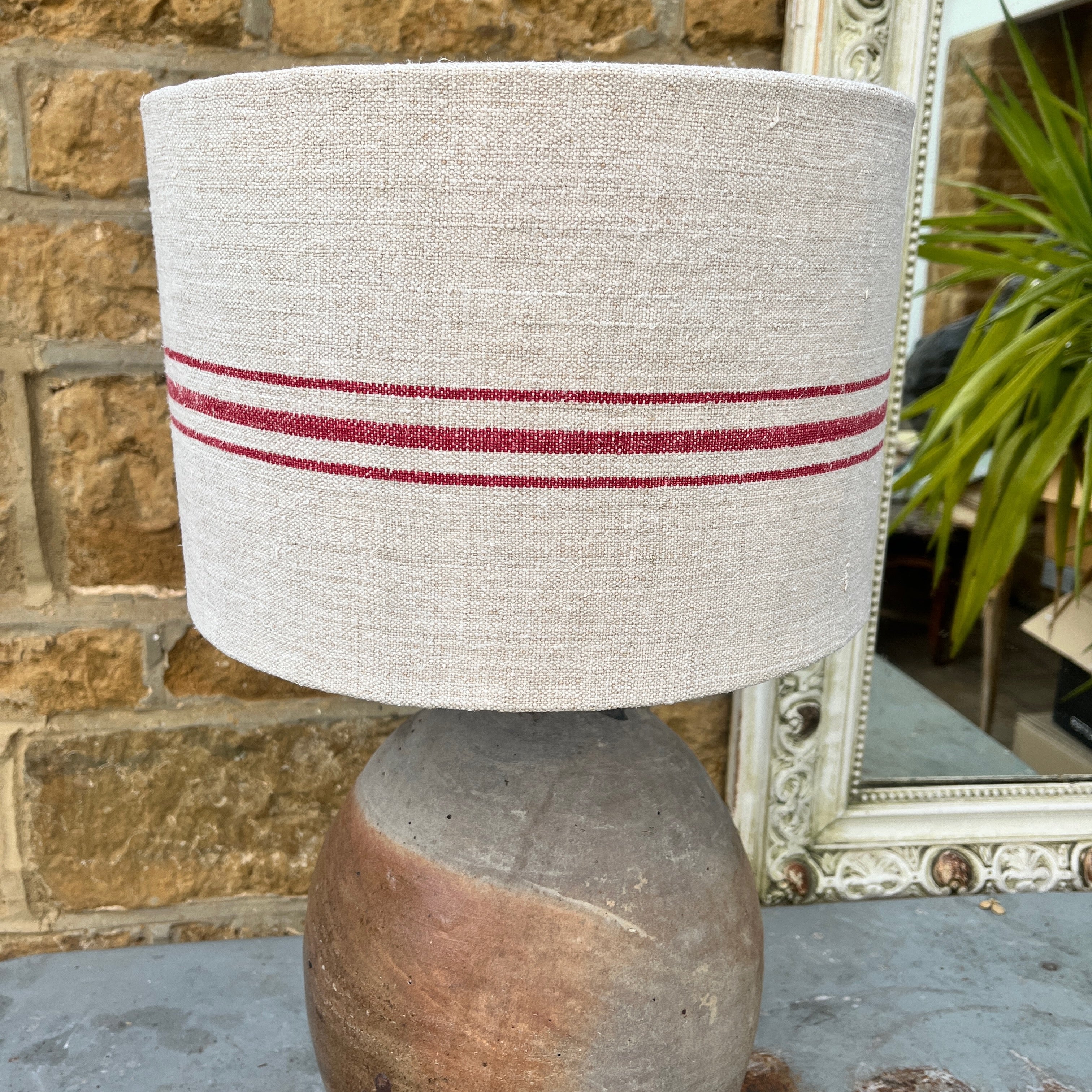A lamp crafted from a repurposed French olive oil jar, featuring a rustic terracotta base with a narrow neck, topped with a handmade shade made from a Hungarian grain sack. The shade showcases a textured, neutral fabric with subtle red stripe around it.