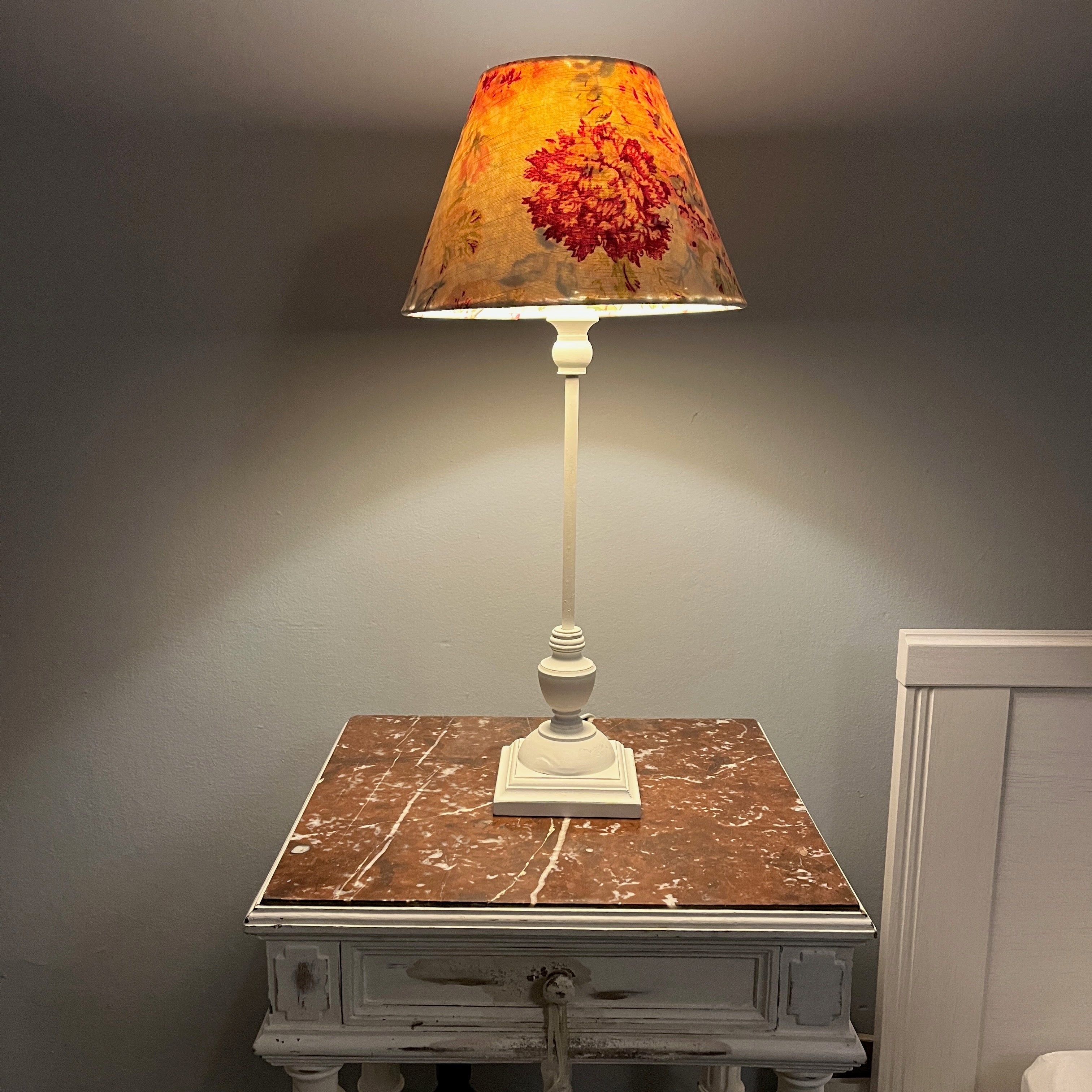 A handmade lampshade made from vintage French linen which sits atop a simple white base. The white base provides a clean, minimalist contrast, highlighting the delicate details of the shade. The overall design evokes a sense of understated sophistication and vintage charm.